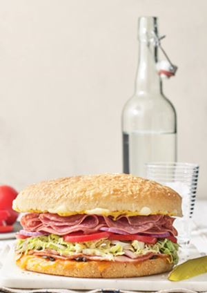 Big sandwich filled with meat and lettuce with a glass bottle filled with water behind it.